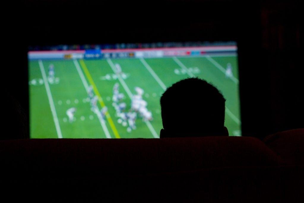 Silhueta de um homem assistindo um jogo de futebol americano na TV.