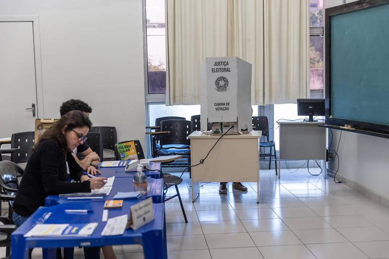 Pessoas sentadas atrás de mesas azuis. Ao fundo, há uma cabina de votação.