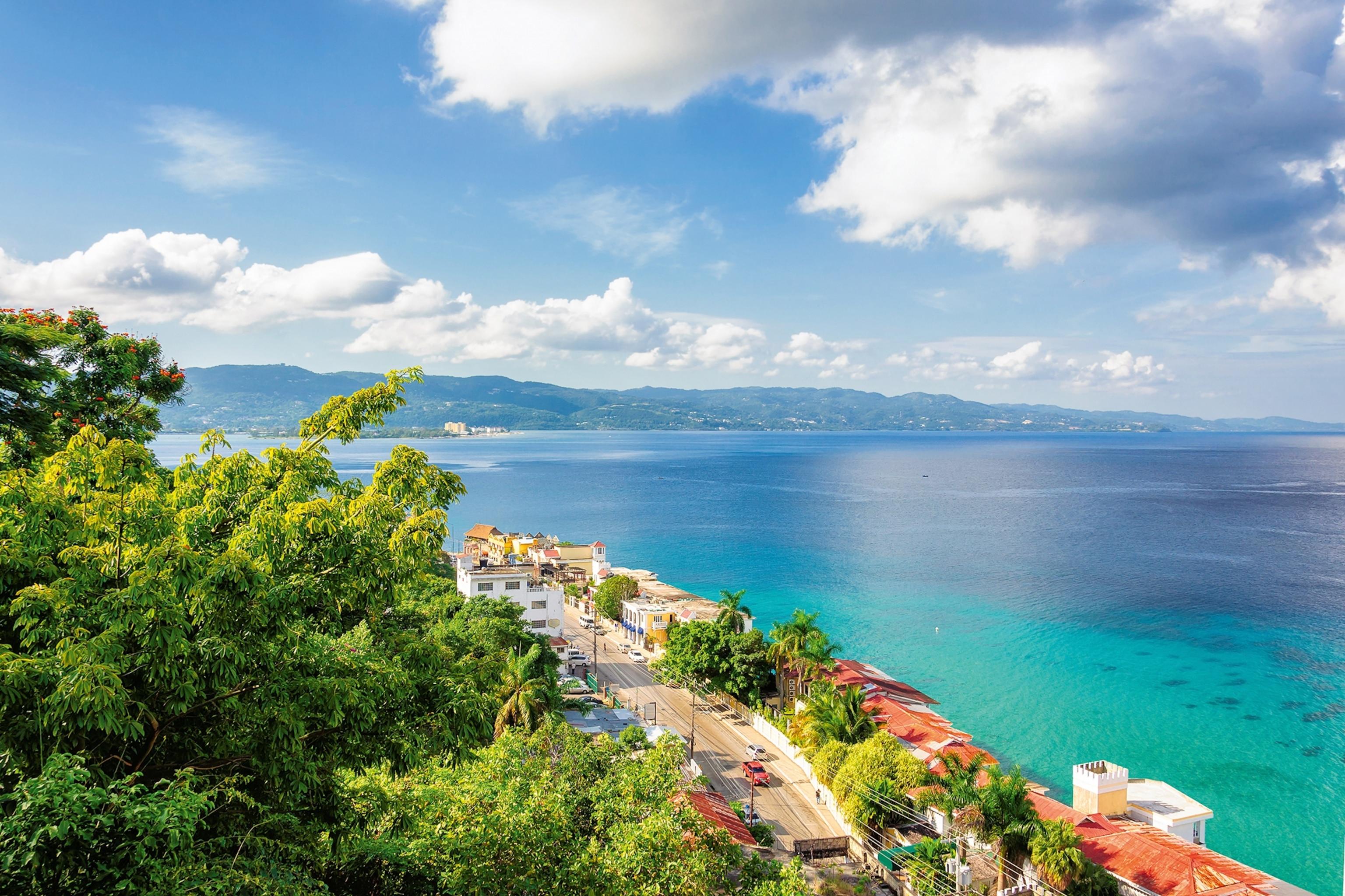 Vista elevada de uma estrada rodeada por vegetação exuberante e edifícios com telhados vermelhos ao lado de um mar turquesa.