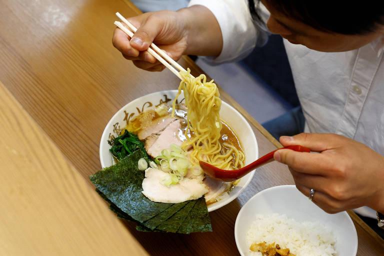 Uma pessoa segurando hashis pegando macarrão de um prato de ramen.