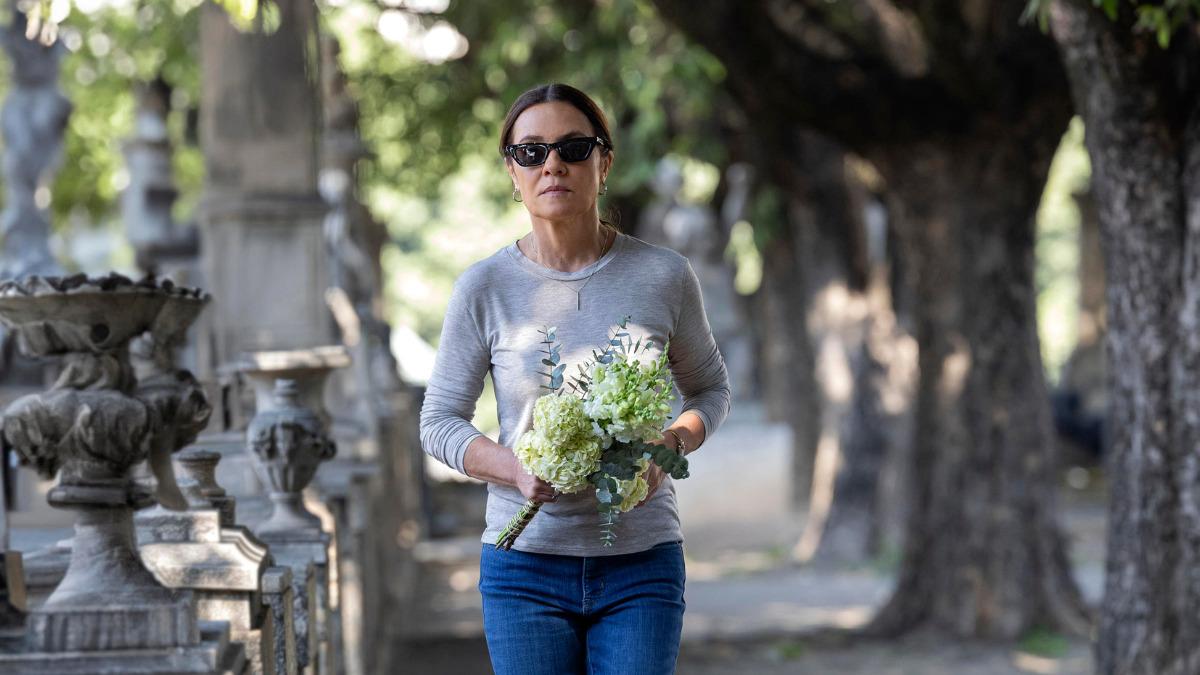 Mércia, personagem de Adriana Esteves, caminha em cemitério usando óculos escuros e segurando um buquê de flores brancas.