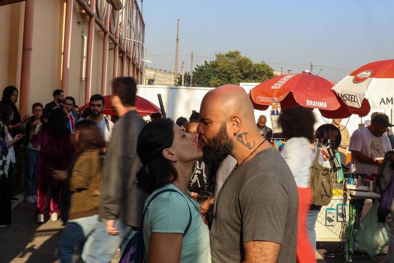 Um casal se beijando em um ambiente movimentado, com várias pessoas ao fundo.