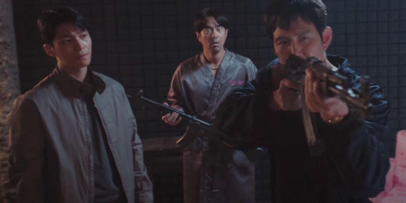 Gi-hun segurando uma arma automática em 'Squid Game' enquanto Jun-ho e Woo-seok reagem ao seu redor.