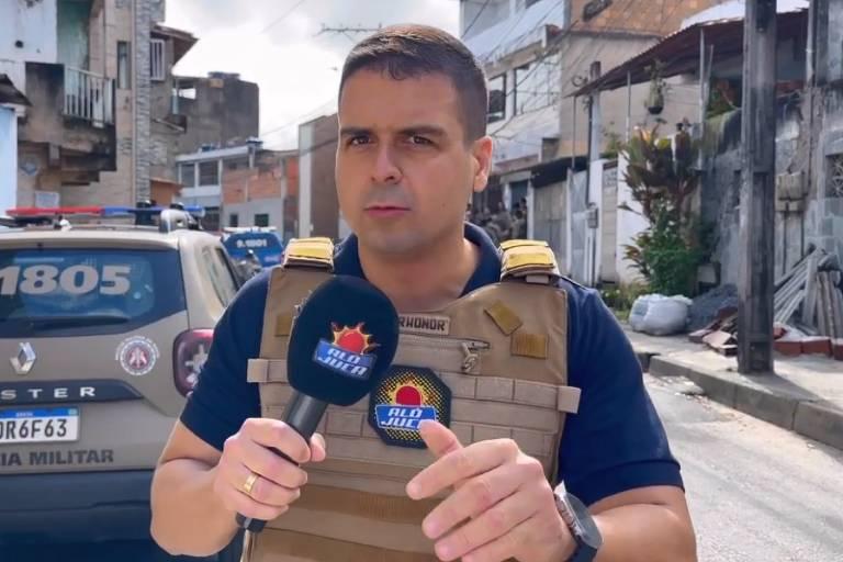 Homem em colete à prova de balas segurando um microfone com viatura de polícia ao fundo.