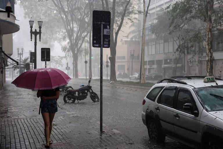 Rua urbana durante uma forte chuva com uma pessoa caminhando sob um guarda-chuva rosa.