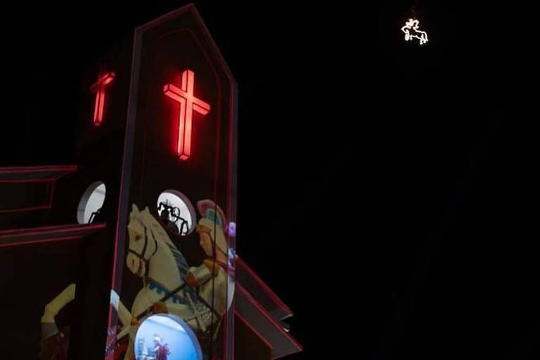 Céu escuro, iluminado por feixes de luz e a projeção da imagem de São Jorge na parede de uma igreja.