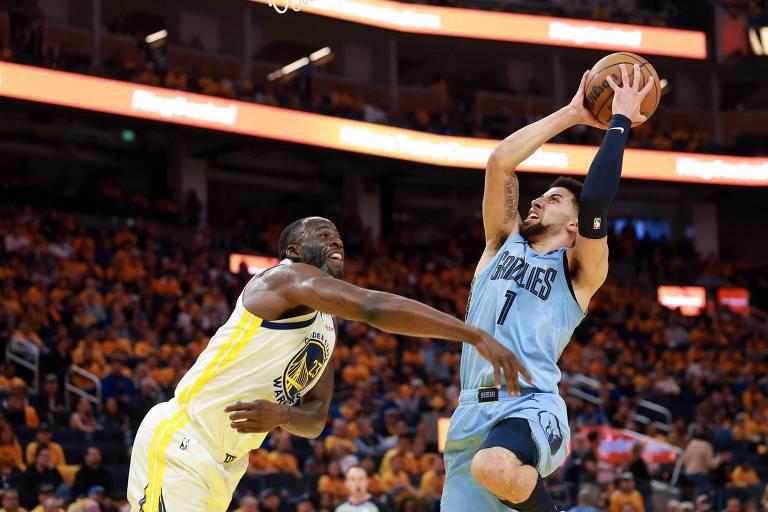 Jogador de basquete saltando para fazer uma cesta, defendido por jogador adversário, em partida de basquete.