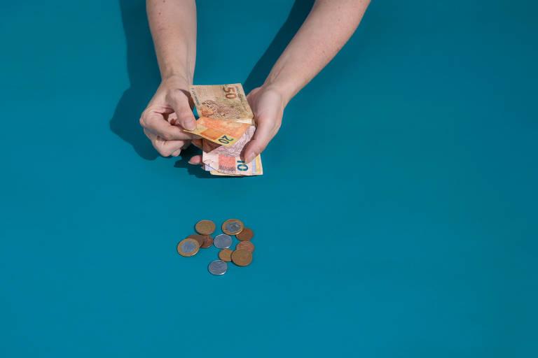 Pessoa segurando notas de dinheiro, com destaque para uma nota de 50 e moedas na mesa.