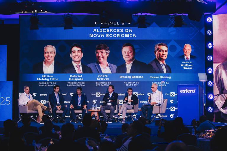 Painel de discussão 'ALICERCES DA NOVA ECONOMIA' com seis homens sentados no palco