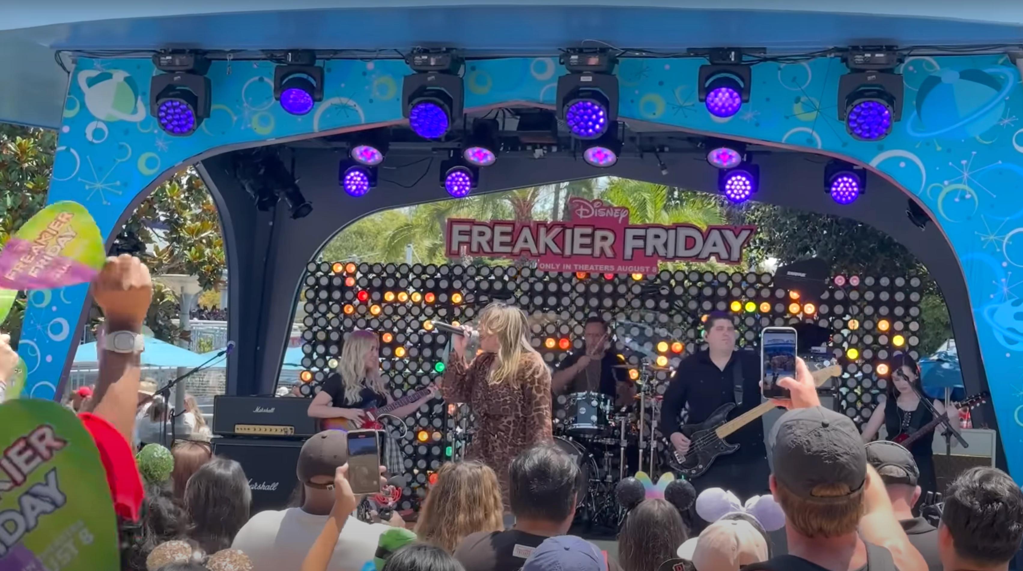 Band performing on stage at Disney's Freakier Friday event with a large crowd watching. A sign displaying the event name is visible in the background.