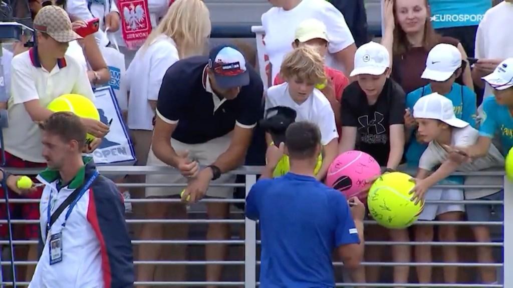 Tennis player signing autographs for young fans after a match. A man steals a cap from one of the boys.