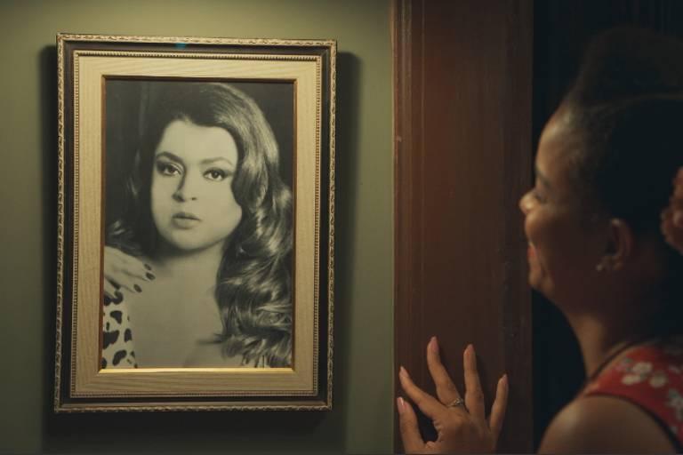 A imagem mostra uma mulher olhando para um retrato em preto e branco de outra mulher, que tem cabelos longos e ondulados