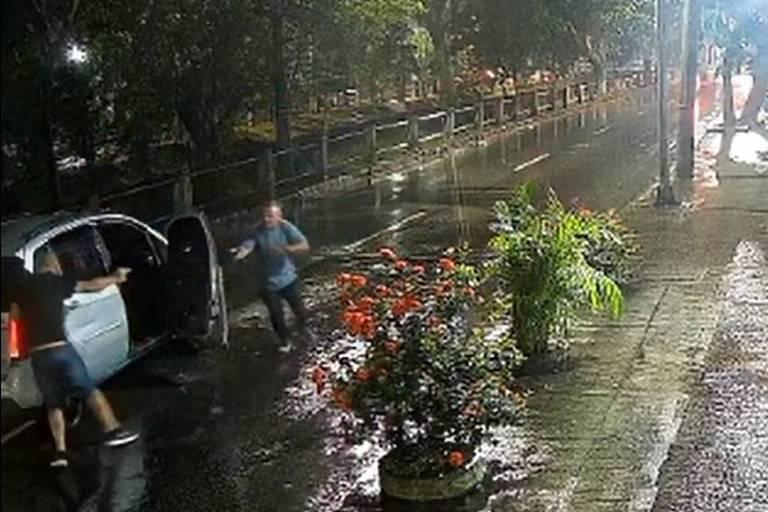 Rua iluminada à noite com calçada molhada e três pessoas próximas a um carro