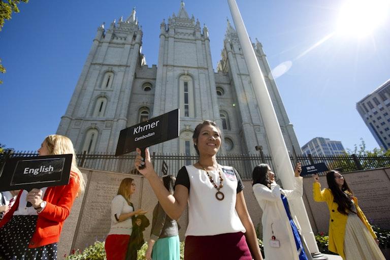 Mulheres seguram pequenos cartazes enquanto estão do lado de fora, em frente a um prédio branco com altas torres.