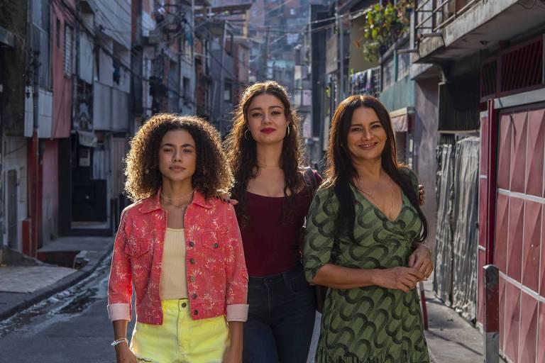 Três mulheres em pé no centro de uma rua estreita de bairro urbano, refletindo a ambientação da nova novela 'Três Graças'.