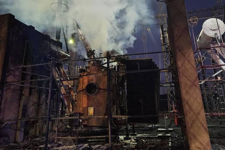 Fumaça densa branca sai de estrutura metálica durante a noite. Ambiente é cercado por grades e postes.