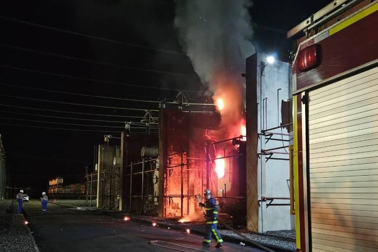 Fogo e fumaça saem de subestação elétrica durante a noite. Bombeiro com mangueira combate as chamas.