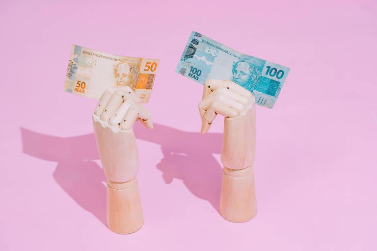 Duas mãos segurando notas de 50 e 100 reais sobre fundo rosa.
