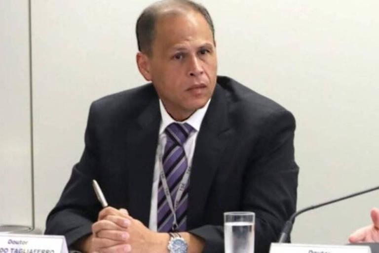 Homem de meia-idade veste terno preto, camisa branca e gravata listrada roxa, sentado à mesa com microfone à esquerda e copo de água à frente. Ambiente interno com parede branca ao fundo.