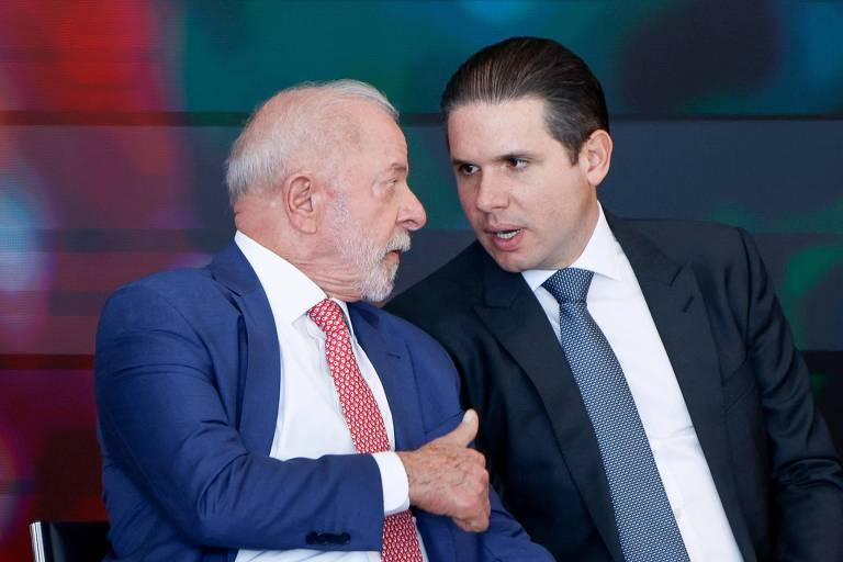 Dois homens vestidos com ternos escuros conversam próximos um do outro. O homem à esquerda tem cabelo branco e barba, veste terno azul e gravata vermelha, enquanto o homem à direita tem cabelo escuro penteado para trás, veste terno preto e gravata azul.