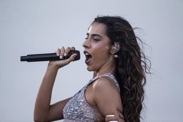 Uma mulher com cabelo longo e ondulado está cantando em um palco. Ela segura um microfone com uma mão e parece estar se expressando intensamente.
