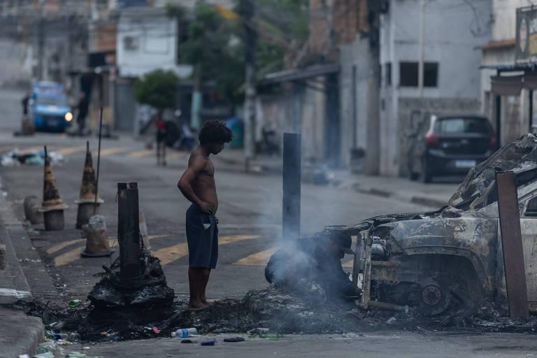Homem observa carro queimado em rua de comunidade urbana