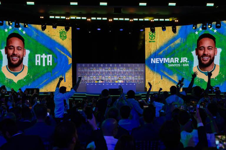 Palco com duas grandes telas exibindo a imagem de Neymar Jr. com a bandeira do Brasil ao fundo. Público em primeiro plano registra o momento com celulares e gestos de comemoração.