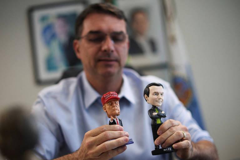 Flávio Bolsonaro segurando bonecos bobblehead de Trump e Jair Bolsonaro