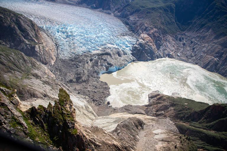 Glaciar azul claro avança entre montanhas rochosas com vegetação esparsa. À frente, lago turvo formado pelo degelo do gelo, cercado por terreno rochoso e encostas verdes.