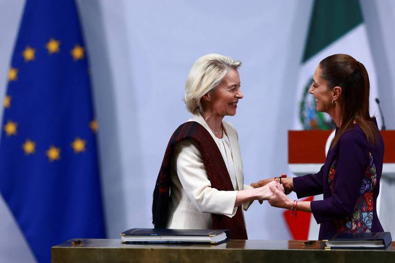 Duas mulheres sorriem e apertam as mãos em frente a uma mesa com documentos, com as bandeiras da União Europeia e do México ao fundo, em ambiente formal.