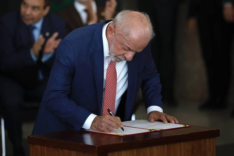 Homem de terno azul e gravata vermelha assina documento sobre mesa de madeira, enquanto quatro pessoas ao fundo aplaudem.