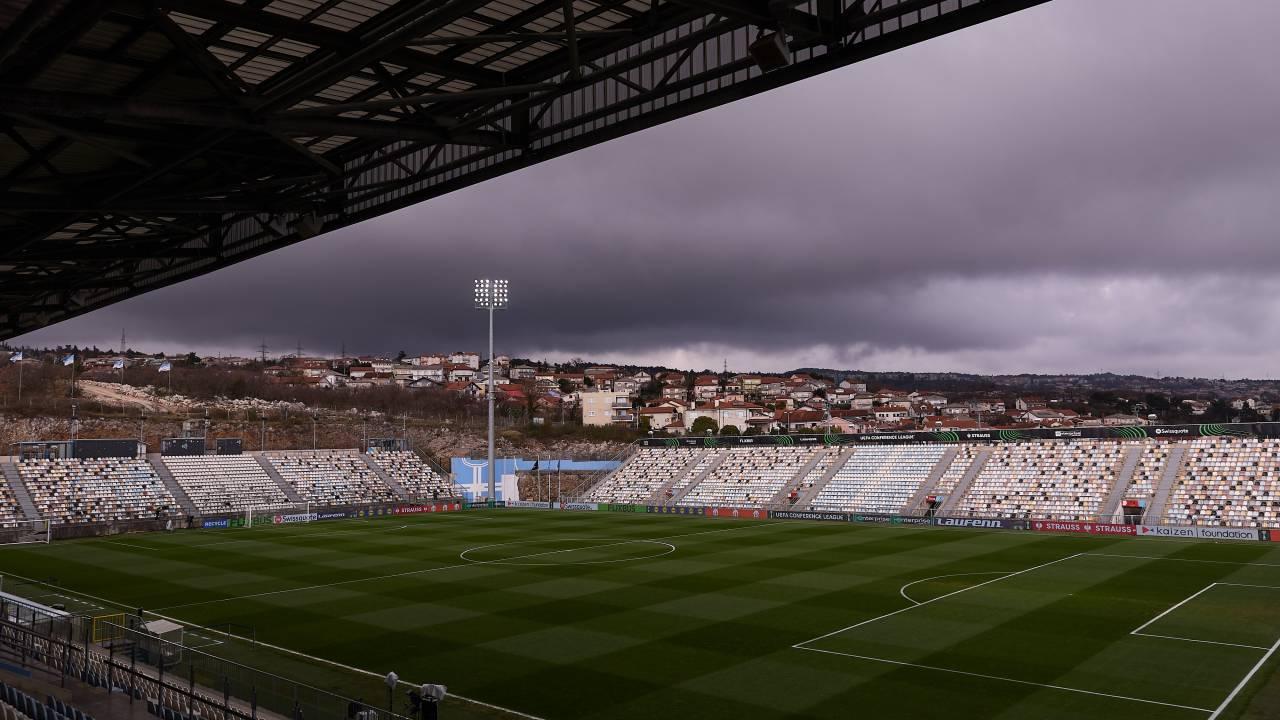 Vista panorâmica de um estádio de futebol vazio, com gramado verde listrado e arquibancadas brancas. O céu está nublado e escuro, com nuvens carregadas.