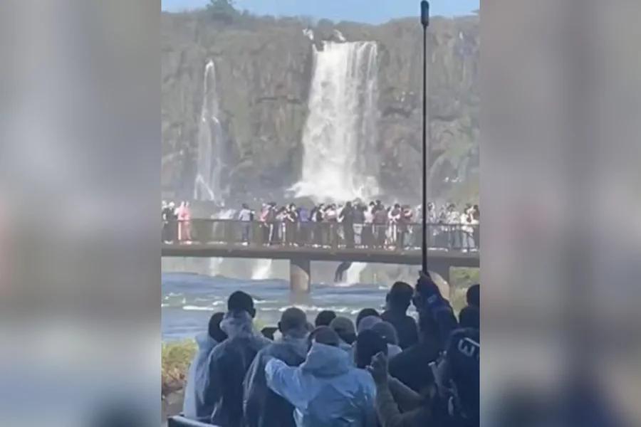 Imagem colorida mostra turista descendo de ponto nas Cataratas do Iguaçu para pegar telefone que havia caído