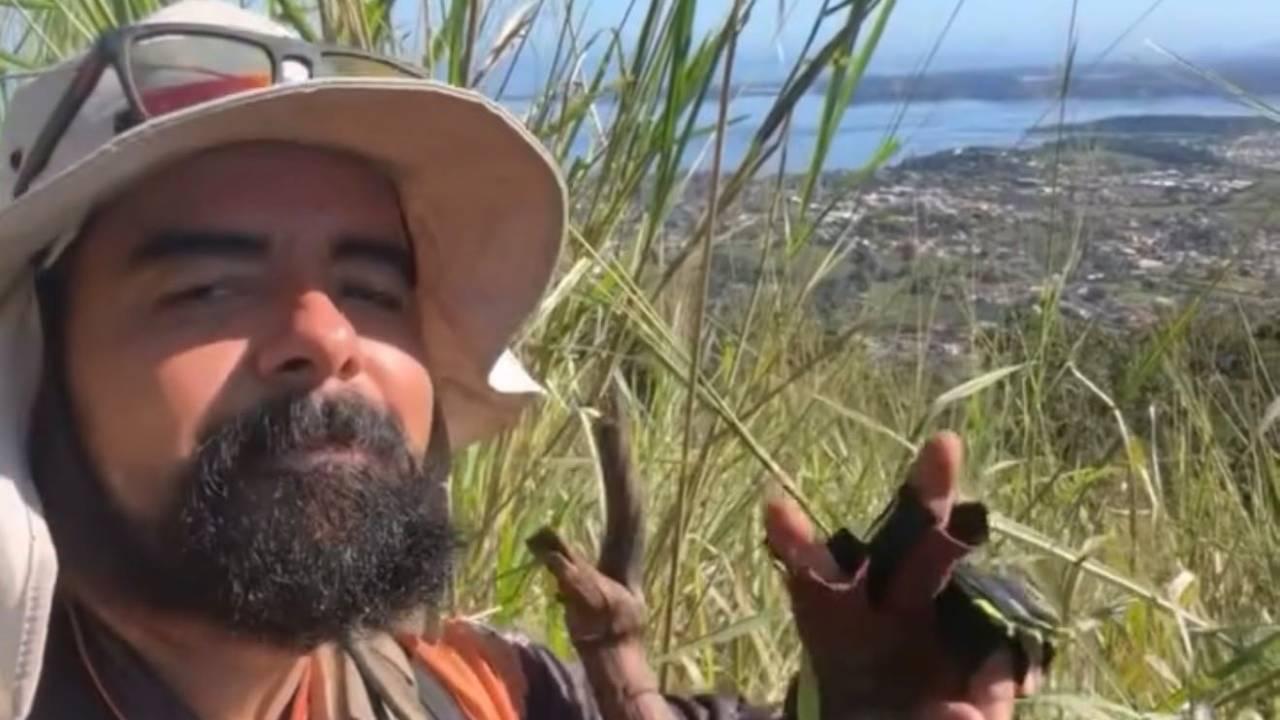 Homem barbudo de chapéu e óculos de sol, sorrindo em área de vegetação, com cidade ao fundo.