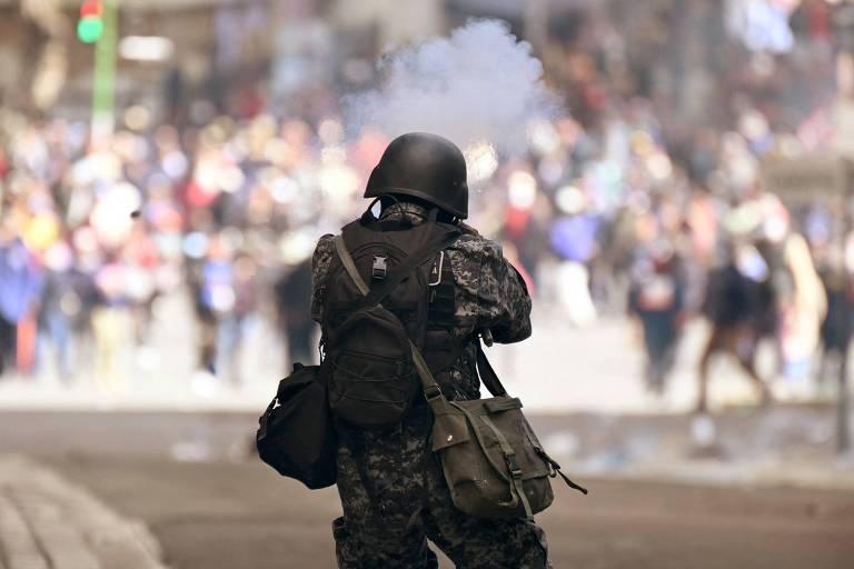 Militar com uniforme camuflado e capacete dispara arma de fogo em direção à multidão.