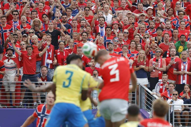Jogadores de futebol do Brasil e da Noruega disputam bola aérea em estádio lotado. Torcida norueguesa observa atentamente.