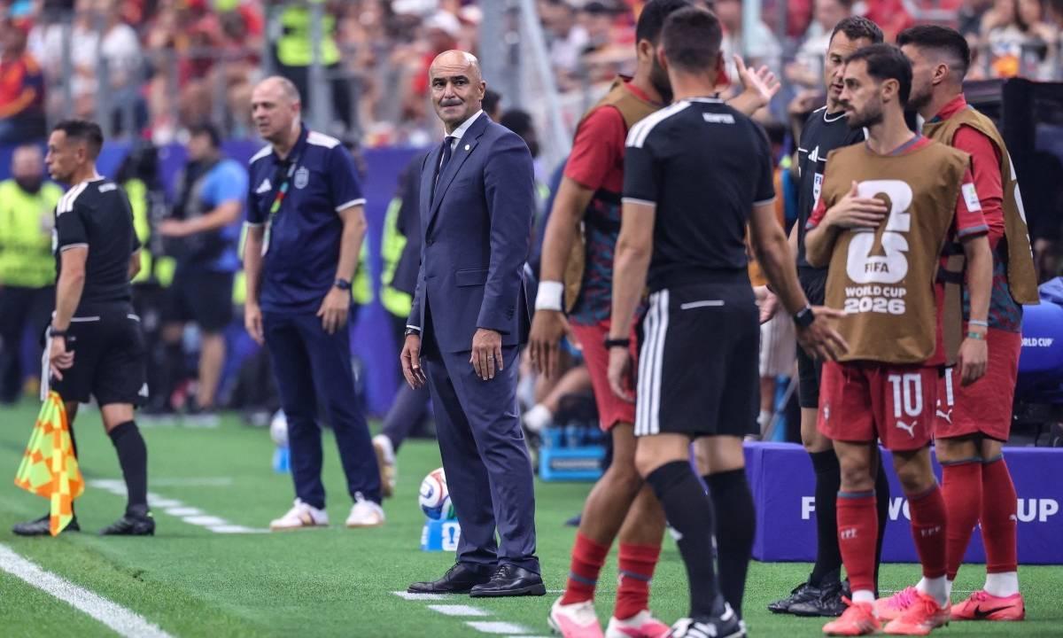 Técnico de terno azul escuro sorri para a câmera no campo de futebol, com jogadores e árbitros ao redor.