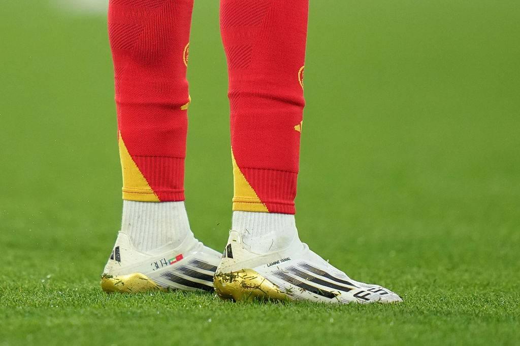 Pés de um jogador de futebol em campo, usando meias vermelhas com detalhes amarelos, meias brancas e chuteiras brancas com solado dourado e listras pretas.