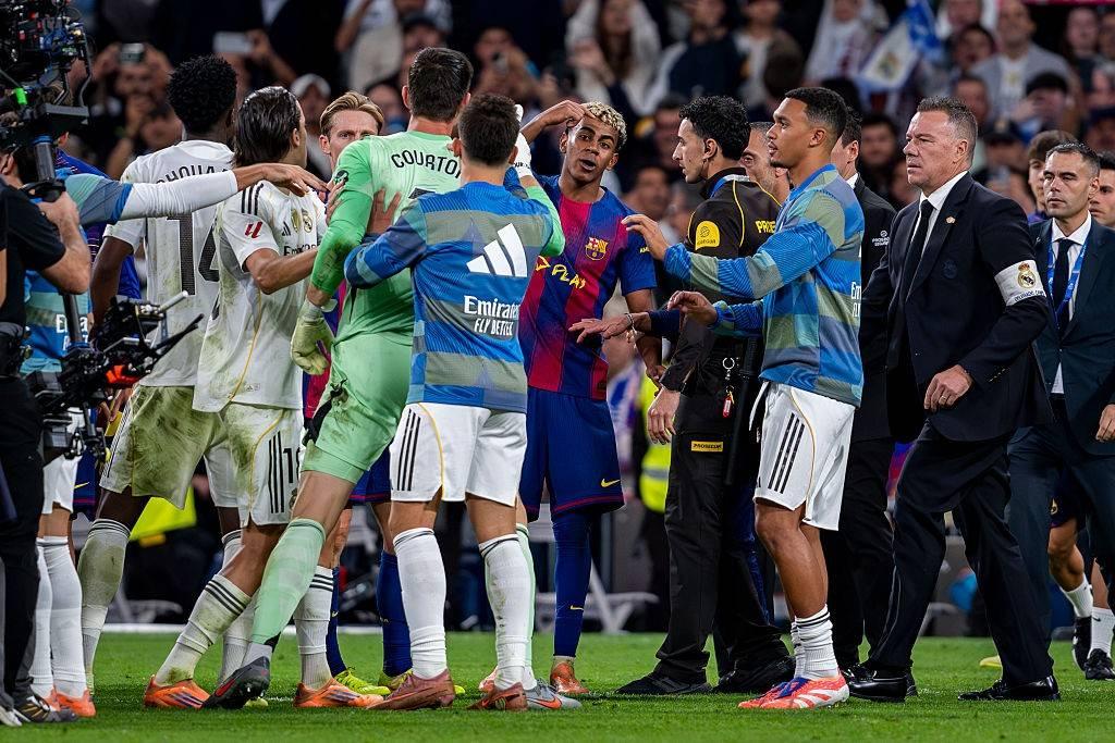 Jogadores de futebol e membros da equipe técnica discutem em campo, com um jogador de camisa azul e vermelha gesticulando no centro, enquanto outros tentam acalmar a situação.