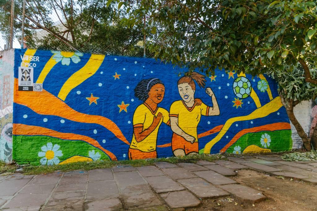 Mural colorido com duas mulheres jogadoras de futebol, uma negra e outra branca, vestindo uniformes amarelos e laranjas, celebrando sob um céu azul com estrelas.