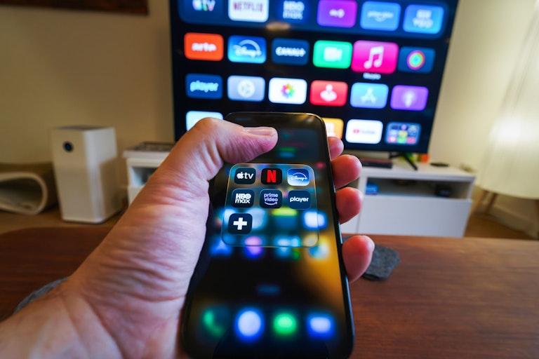 Uma TV em segundo plano enquanto alguém segura o celular com aplicativos de streaming à frente.