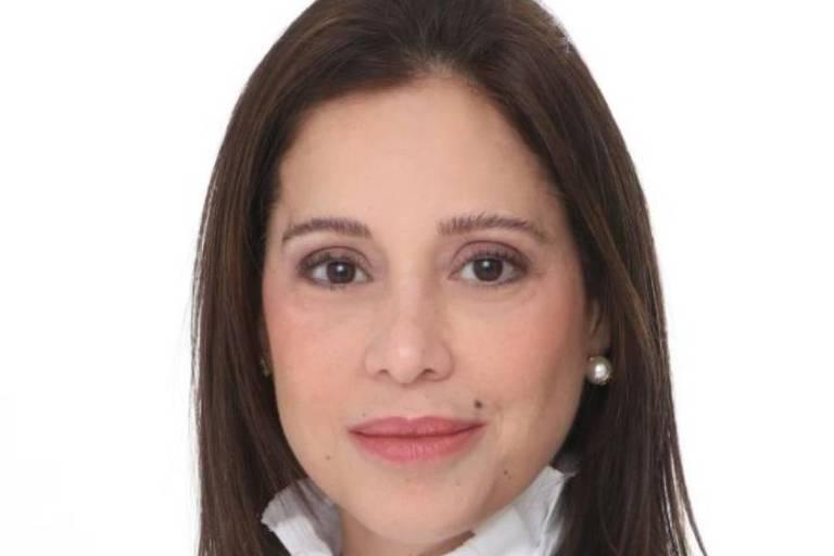 Mulher de cabelos castanhos lisos, usando blusa branca com babados e blazer preto, posando contra fundo branco.